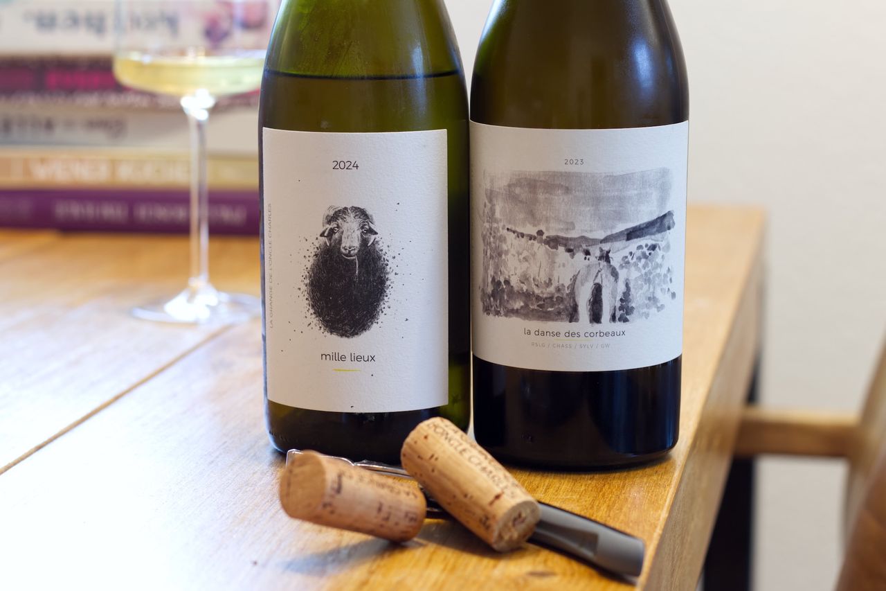 Auf einem Holztisch stehen zwei Flaschen Wein vom Weingut La Grange de l'Oncle Charles. Im Hintergrund sind ein Weinglas und ein Bücherstapel zu sehen. Vor den Flaschen liegt ein Korken am Kellnermesser.