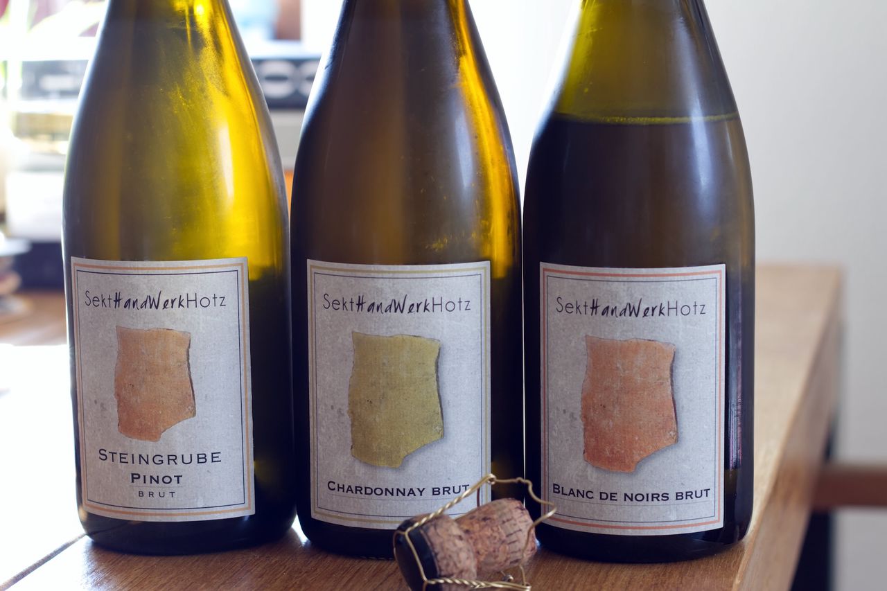 Auf einem Holztisch stehen zwei Flaschen Sekt vom Sekthandwerk Hotz. Im Hintergrund sind ein Weinglas und ein Bücherstapel zu sehen. Vor den Flaschen liegt ein Korken.