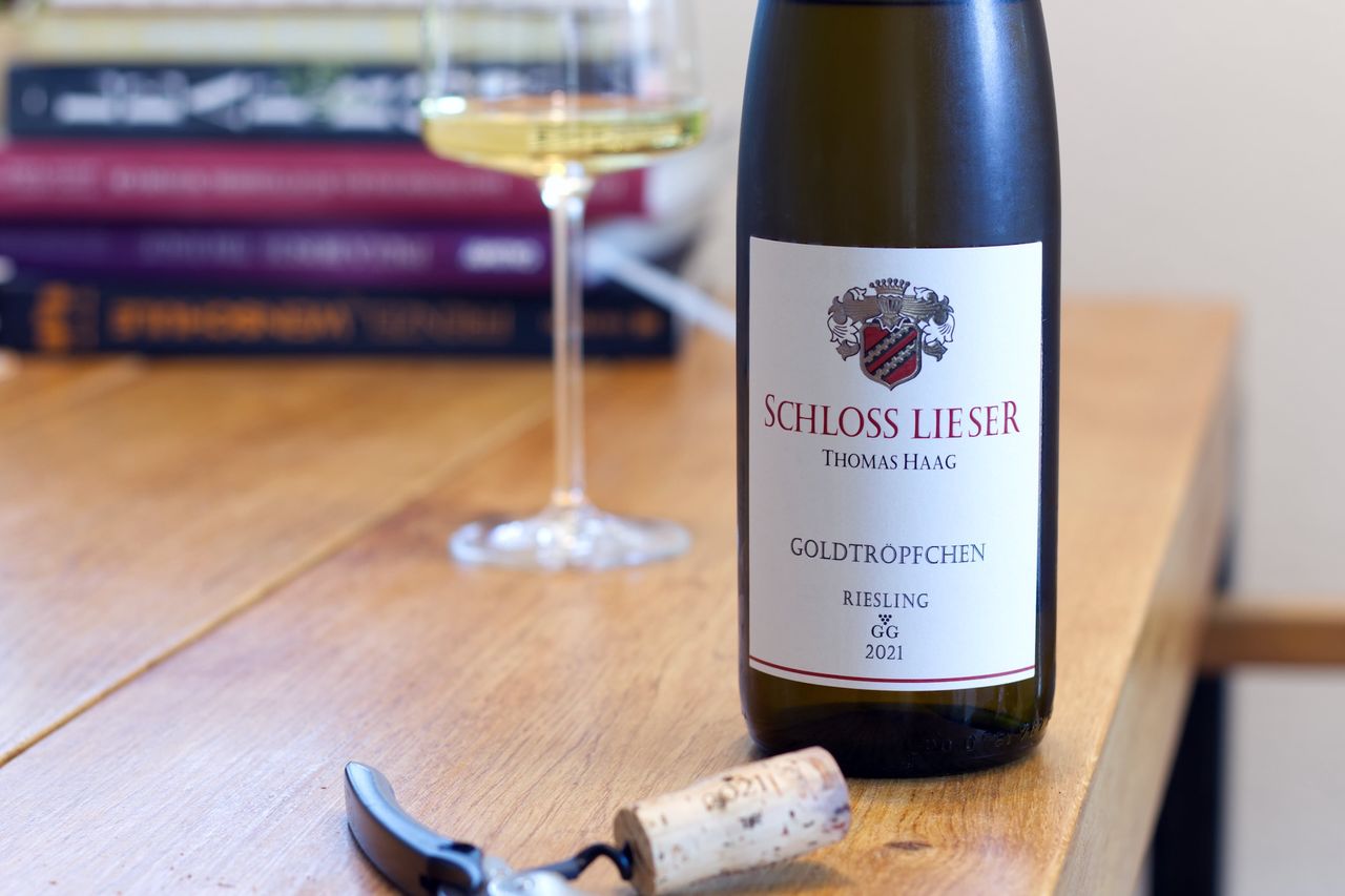 Auf einem Holztisch steht eine Flasche Goldtröpchen Riesling von Schloss Lieser. Im Hintergrund sind ein Weinglas und ein Bücherstapel zu sehen, vor der Flasche liegen Korken und Kellnermesser.