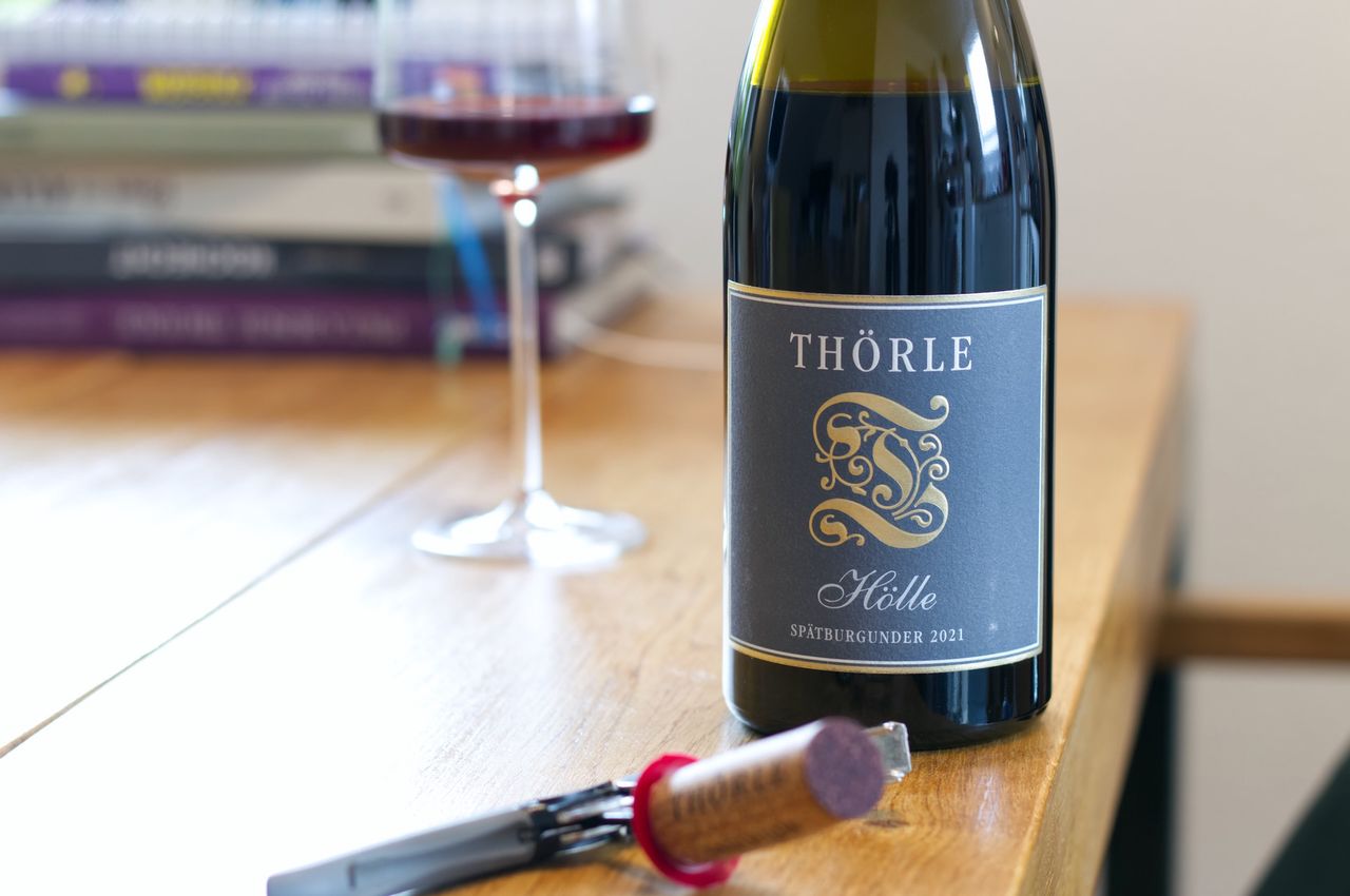 Auf einem Holztisch steht eine Flasche Hölle Spätburgunder vom Weingut Thörle. Im Hintergrund sind ein Weinglas und ein Bücherstapel zu sehen, vor der Flasche liegen Korken und Kellnermesser.
