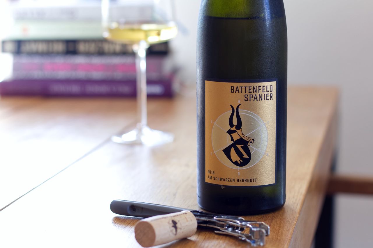 Auf einem Holztisch steht eine Flasche Am Schwarzen Herrgott von Battenfeld-Spanier. Im Hintergrund sind ein Weinglas und ein Bücherstapel zu sehen, vor der Flasche liegen Korken und Kellnermesser.