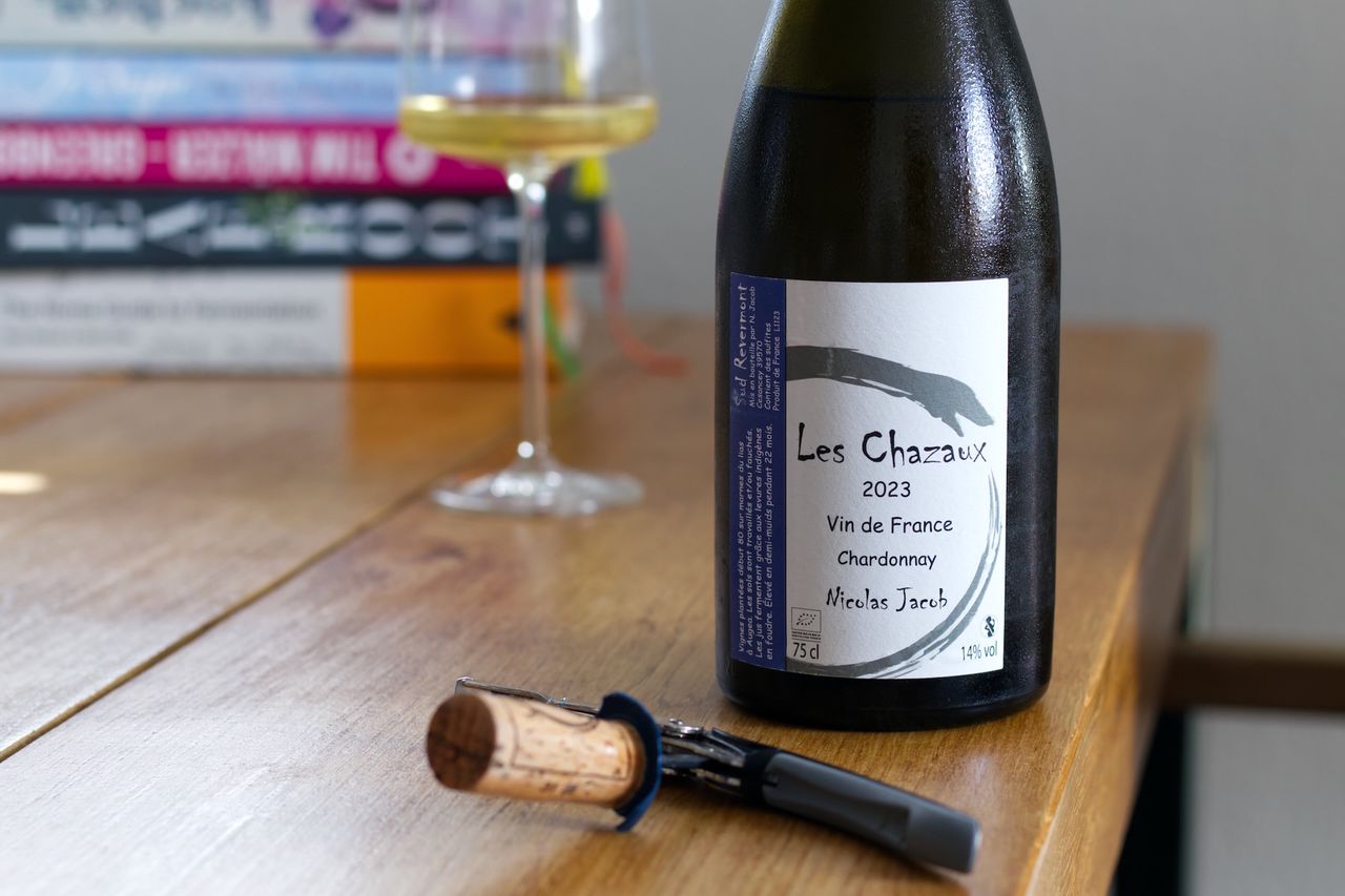 Auf einem Holztisch steht eine Flasche Les Chazaux von Nicolas Jacob. Im Hintergrund sind ein Weinglas und ein Bücherstapel zu sehen, vor der Flasche liegen Korken und Kellnermesser.