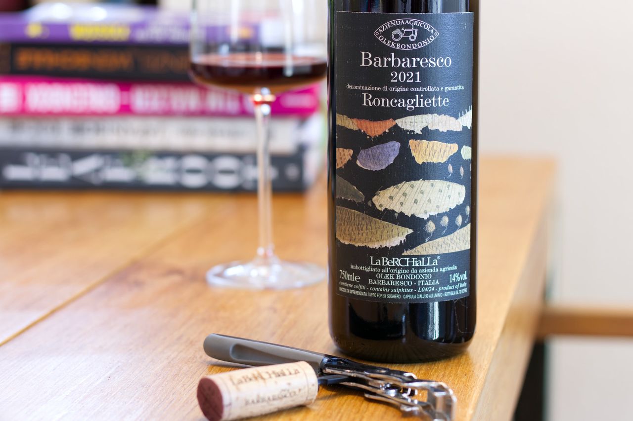 Auf einem Holztisch steht eine Flasche Barbaresco Roncagliette von Olek Bondonio. Im Hintergrund sind ein Weinglas und ein Bücherstapel zu sehen, vor der Flasche liegen Korken und Kellnermesser.