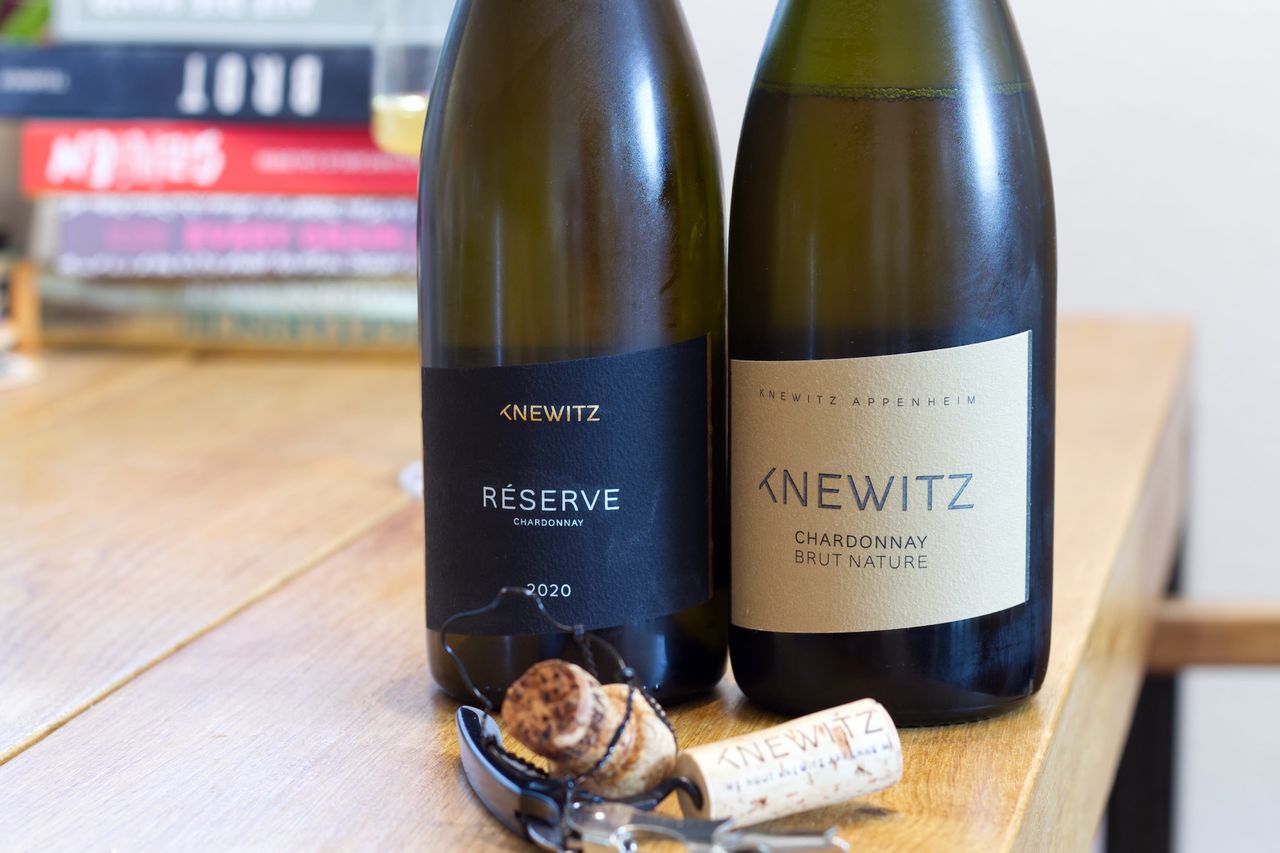 Auf einem Holztisch stehen zwei Flaschen Wein von Knewitz. Im Hintergrund sind ein Weinglas und ein Bücherstapel zu sehen, vor den Flaschen liegen Korken und Kellnermesser.