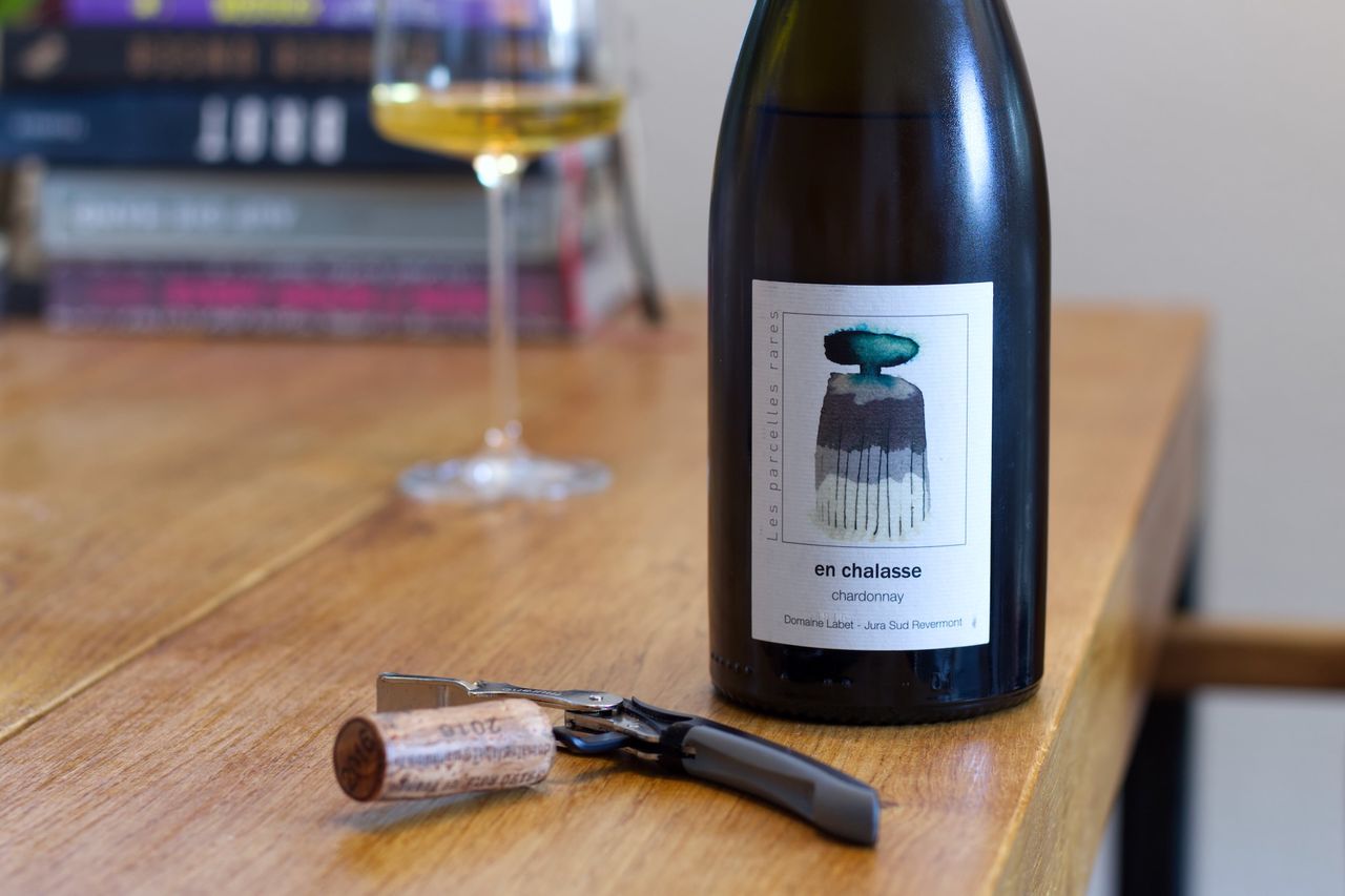 Auf einem Holztisch steht eine Flasche En Chalasse von Labet. Im Hintergrund sind ein Weinglas und ein Bücherstapel zu sehen, vor der Flasche liegen Korken und Kellnermesser.