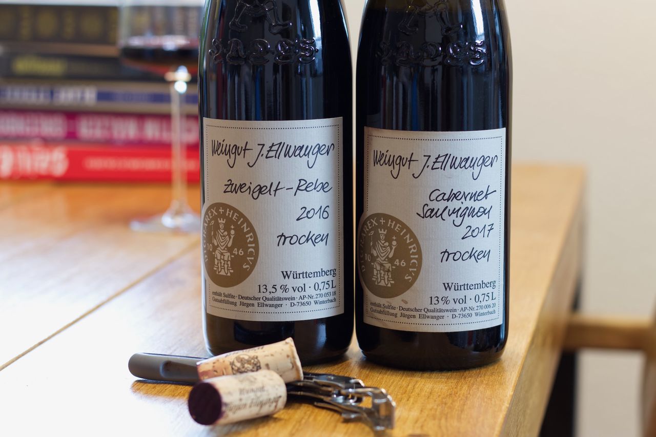 Auf einem Holztisch stehen zwei Flaschen Wein von J. Ellwanger mit dem HADES Logo. Im Hintergrund sind ein Weinglas und ein Bücherstapel zu sehen, vor den Flaschen liegen Korken und Kellnermesser.