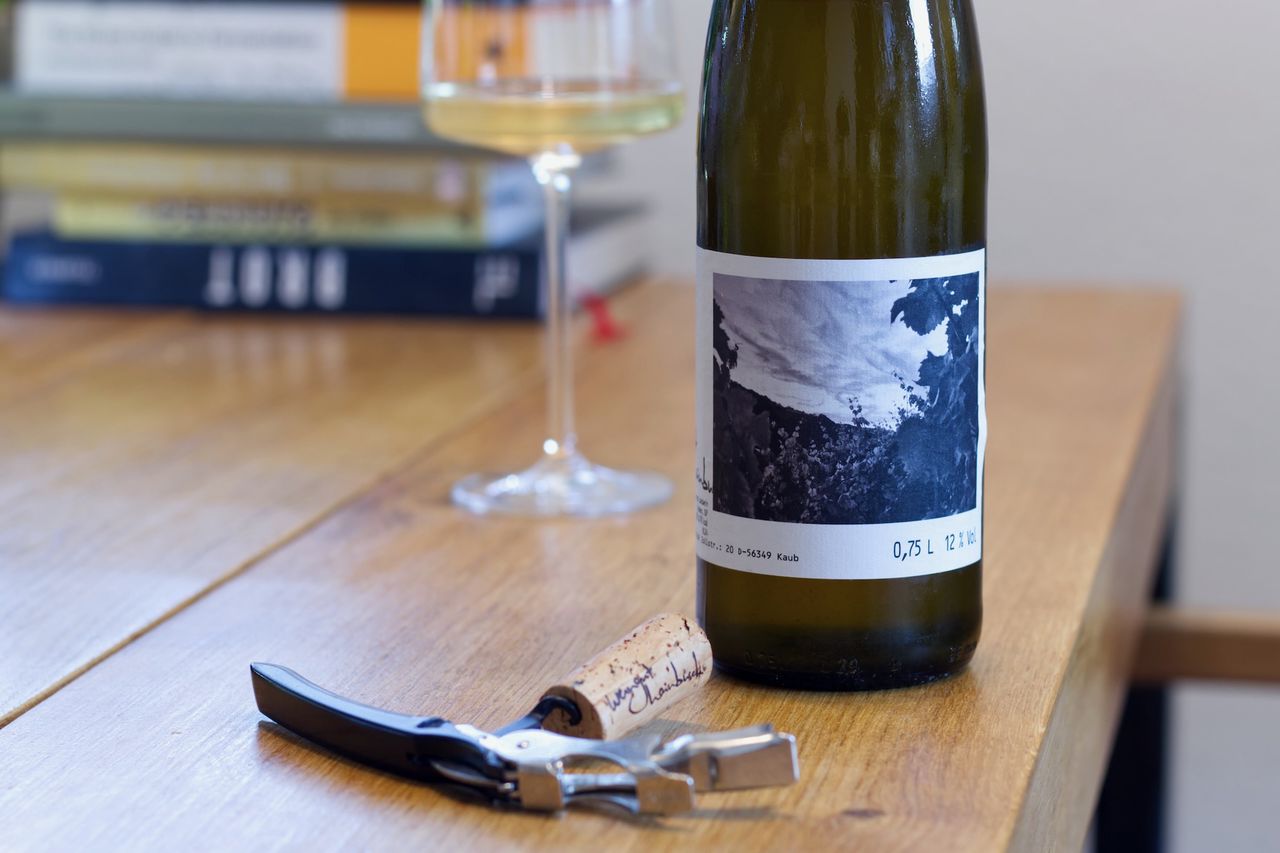 Auf einem Holztisch steht eine Flasche Riesling vom Weingut Hainbusch. Im Hintergrund sind ein Weinglas und ein Bücherstapel zu sehen, vor der Flasche liegt Korken und Kellnermesser.