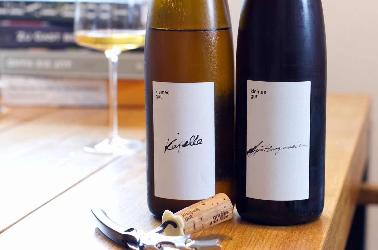 Auf einem Holztisch stehen zwei Flaschen Wein vom kleinen Gut: Ein Chardonnay und ein Spätburgunder. Im Hintergrund sind ein Weinglas und ein Bücherstapel zu sehen, vor den Flaschen liegen Korken und Kellnermesser.
