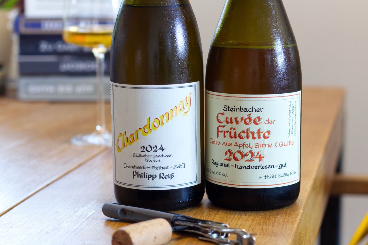 Auf einem Holztisch stehen zwei Flaschen von Philipp Reiß mit Kallifgrafie auf dem Etikett: Ein Chardonnay und eine Flasche Cuvée der Früchte. Im Hintergrund sind ein Weinglas und ein Bücherstapel zu sehen, vor den Flaschen liegen Korken und Kellnermesser.