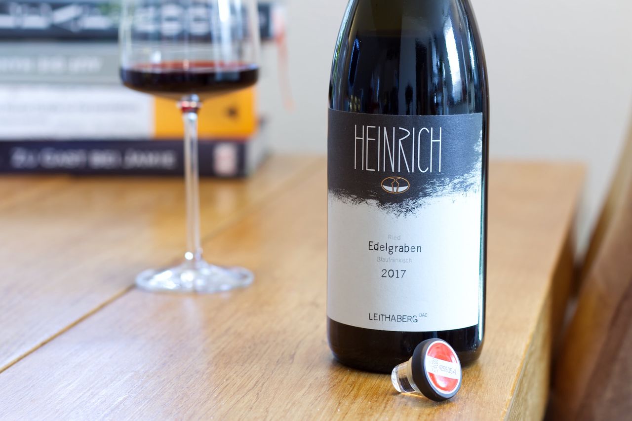Auf einem Holztisch steht eine Flasche Ried Edelgraben von Heinrich. Im Hintergrund sind ein Weinglas und ein Bücherstapel zu sehen, vor der Flasche liegt der Glasverschluss.