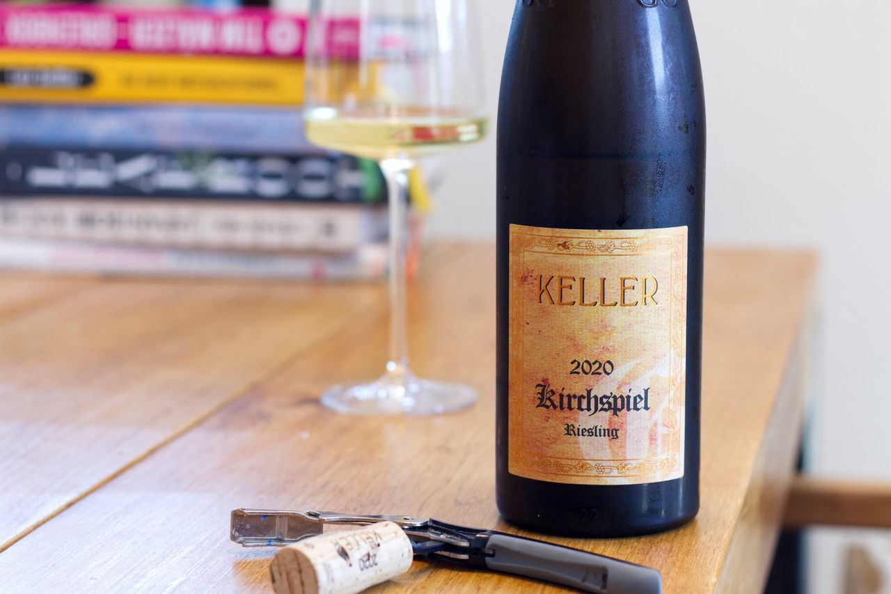 Auf einem Holztisch steht eine Flasche Kirchspiel von Keller. Im Hintergrund sind ein Weinglas und ein Bücherstapel zu sehen, vor der Flasche liegen Korken und Kellnermesser.