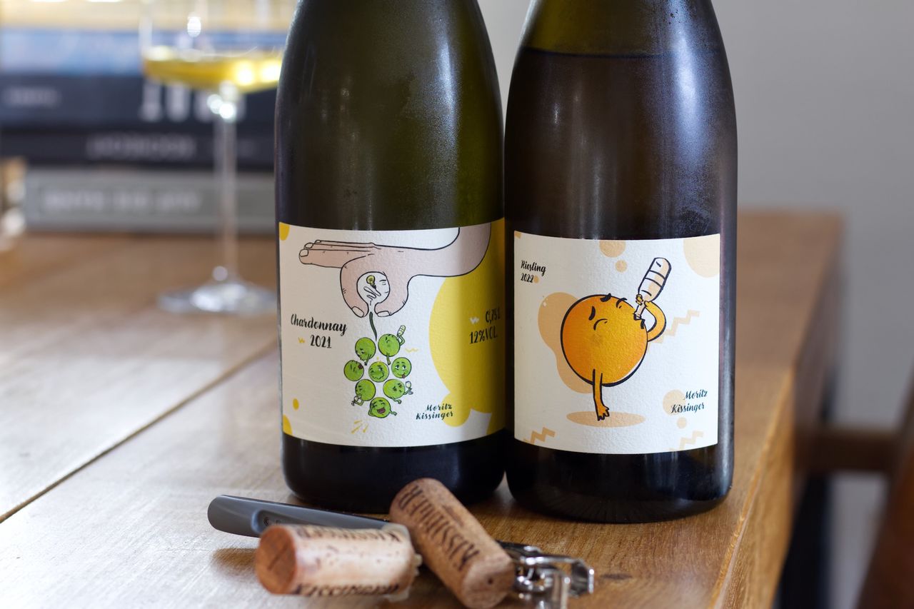Auf einem Holztisch stehen zwei Flaschen Wein mit den Trauben-Etiketten von Moritz Kissinger. Im Hintergrund sind ein Weinglas und ein Bücherstapel zu sehen, vor der Flasche liegen Korken und Kellnermesser.