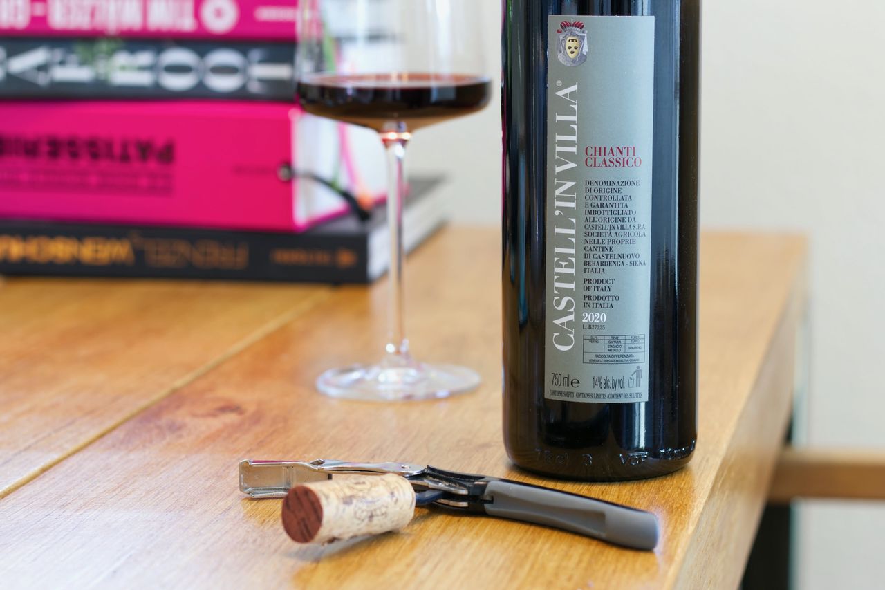 Auf einem Holztisch steht eine Flasche Chianti Classico. Im Hintergrund sind ein Weinglas und ein Bücherstapel zu sehen, vor der Flasche liegen Korken und Kellnermesser.