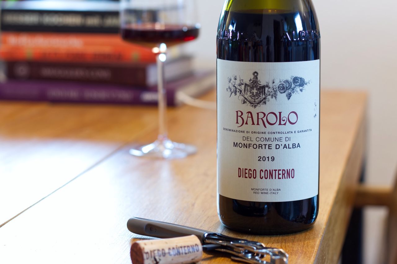 Auf einem Holztisch steht eine Flasche Barolo von Diego Conterno. Im Hintergrund sind ein Weinglas und ein Bücherstapel zu sehen, vor der Flasche liegen Korken und Kellnermesser.