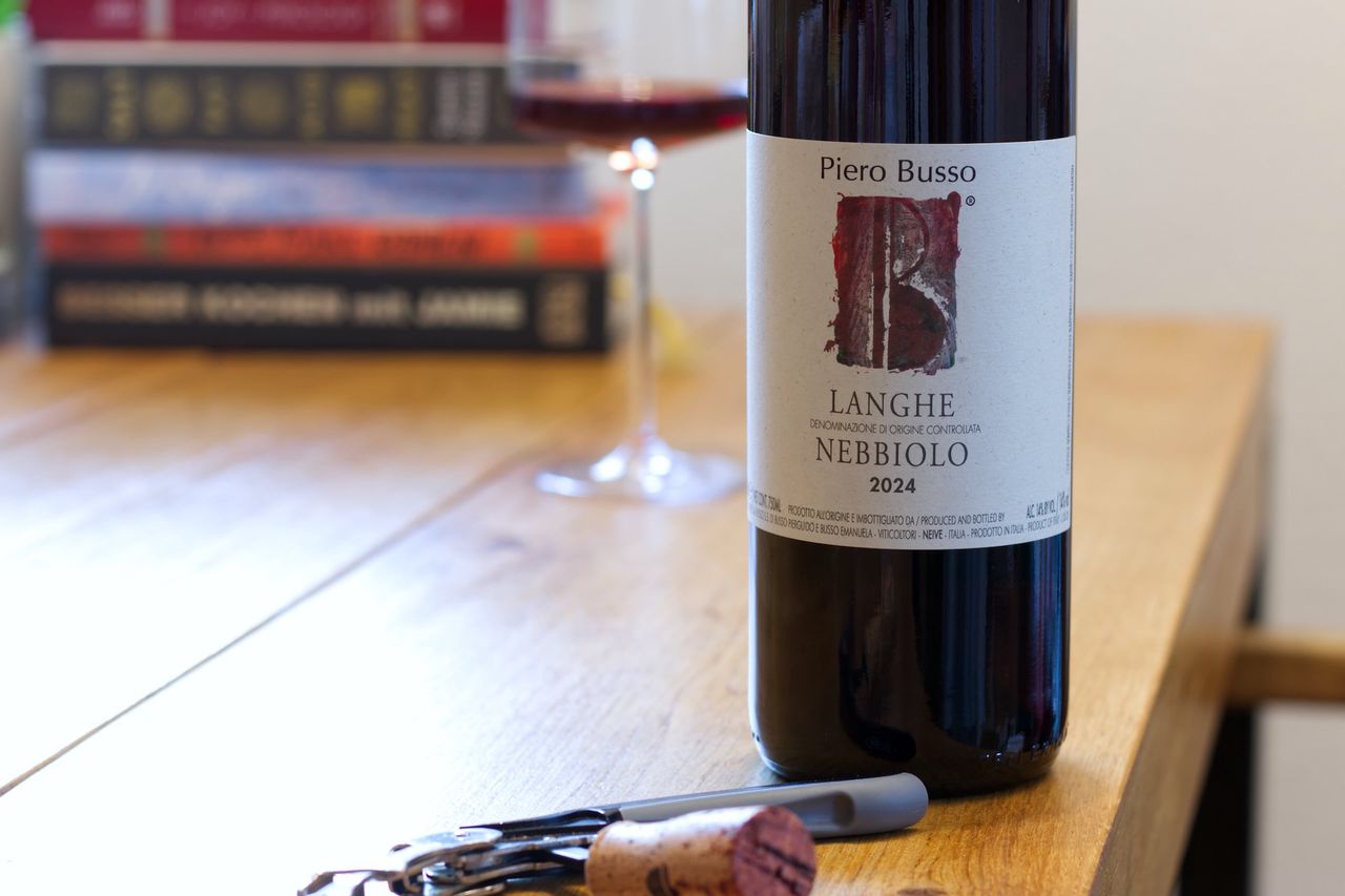 Auf einem Holztisch steht eine Flasche Nebbiolo von Piero Busso. Im Hintergrund sind ein Weinglas und ein Bücherstapel zu sehen, vor der Flasche liegen Korken und Kellnermesser.