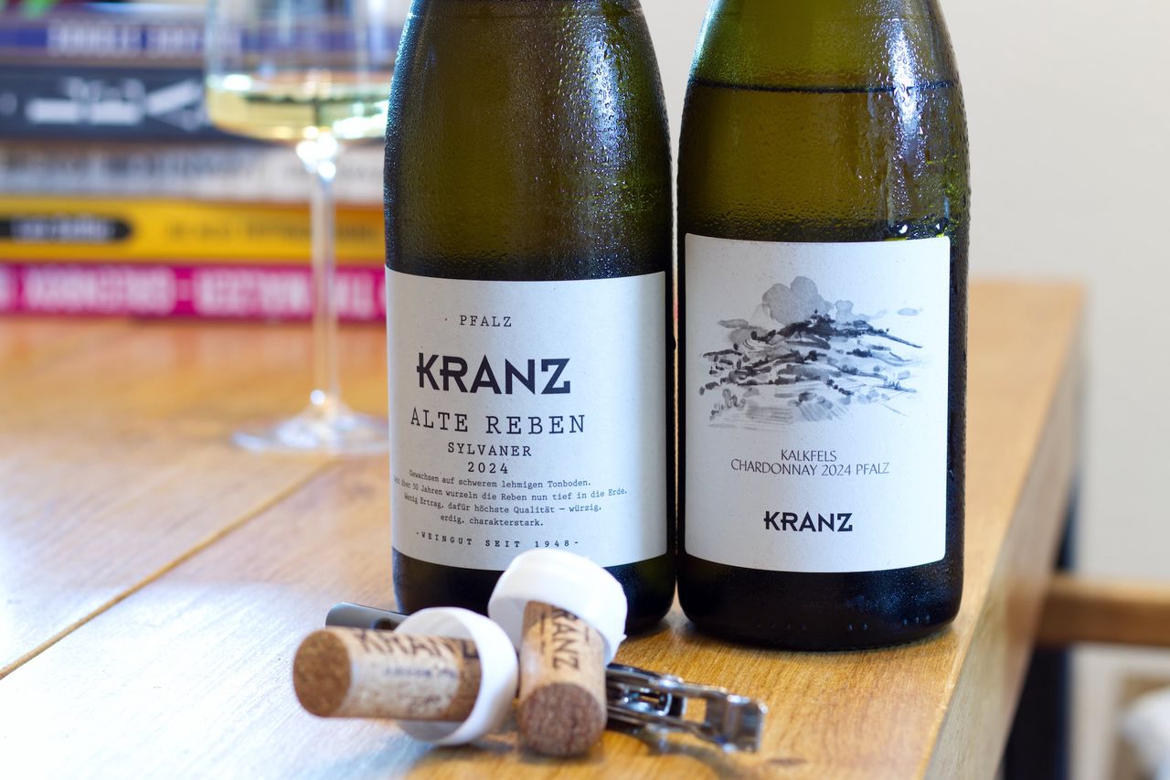 Auf einem Holztisch stehen zwei Flaschen Wein vom Weingut Kranz. Im Hintergrund sind ein Weinglas und ein Bücherstapel zu sehen, vor der Flasche liegen Korken und Kellnermesser.